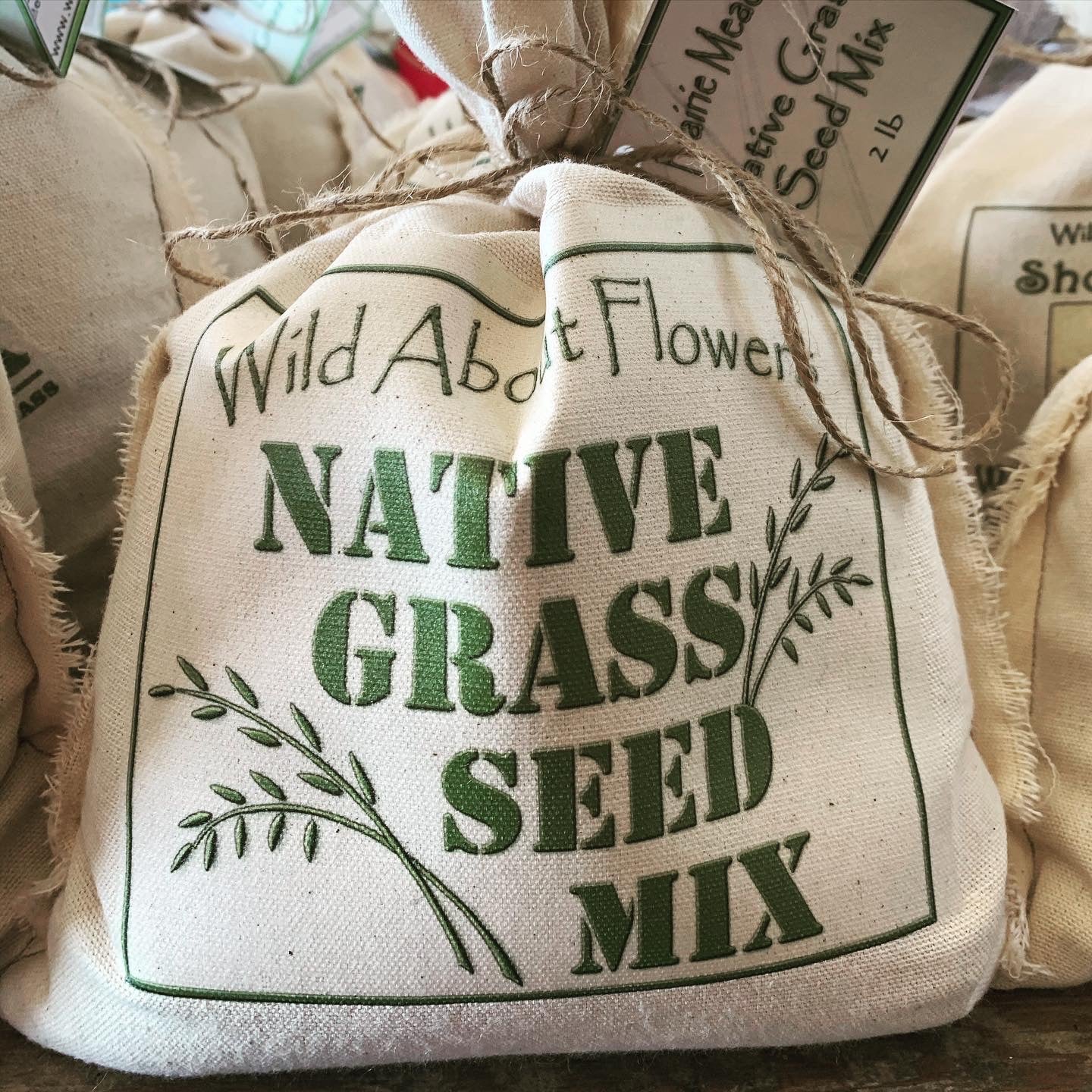 A cloth bag labeled "Wild About Flowers - Native Grass Seed Mix" sits prominently in the foreground, tied at the top with twine. The green lettering is framed by simple illustrations of grass stems. Additional seed bags are visible in the background, contributing to a rustic and natural display of grass seed packaging.
