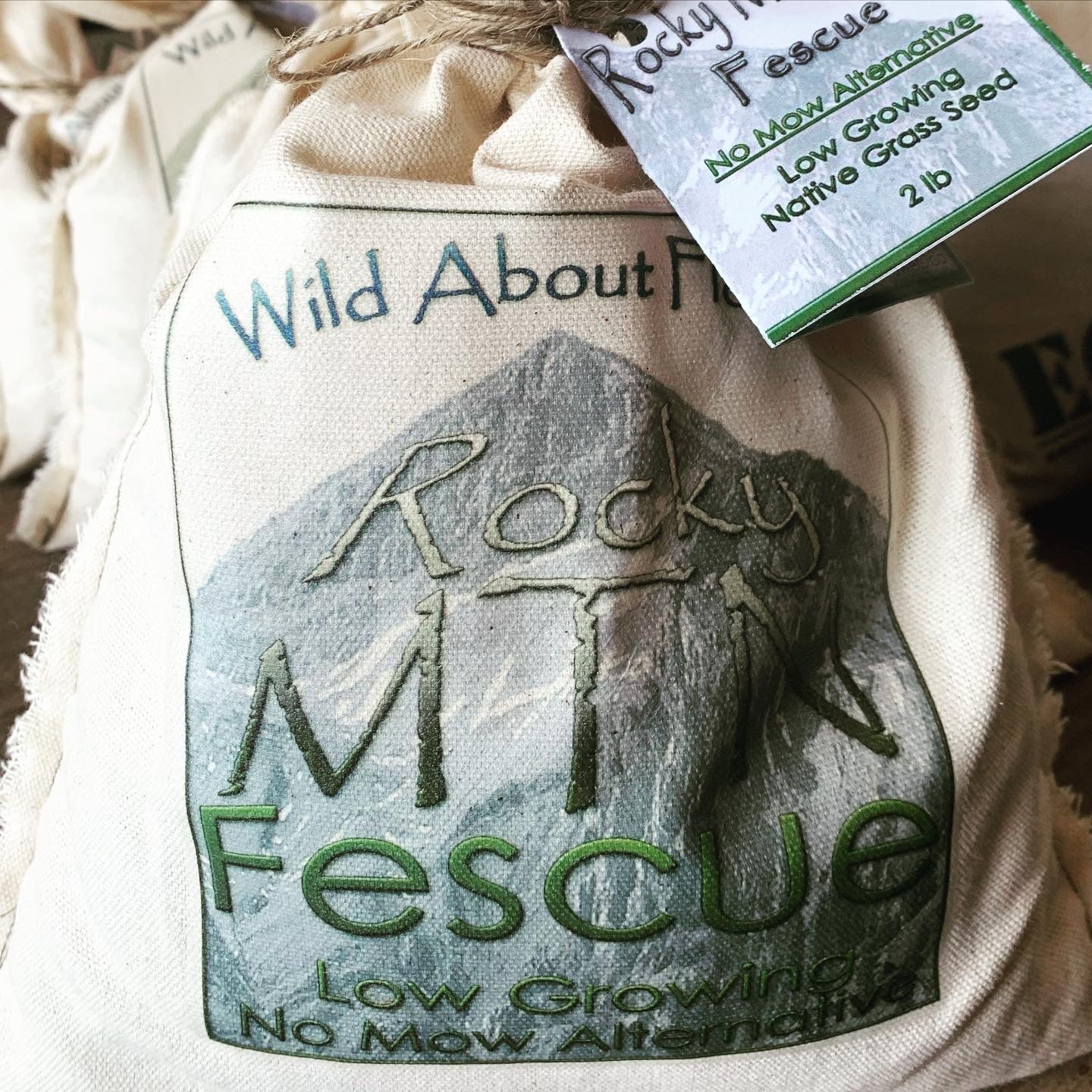 A cloth bag labeled "Wild About Flowers - Rocky MTN Fescue" is featured in the image, tied at the top with twine and a product tag. The front design displays a mountain graphic with text indicating it is a "Low Growing, No Mow Alternative." The tag reads "Rocky Mountain Fescue - No Mow Alternative - Low Growing Native Grass Seed - 2 lb." Other seed bags are partially visible in the background, creating a rustic display.