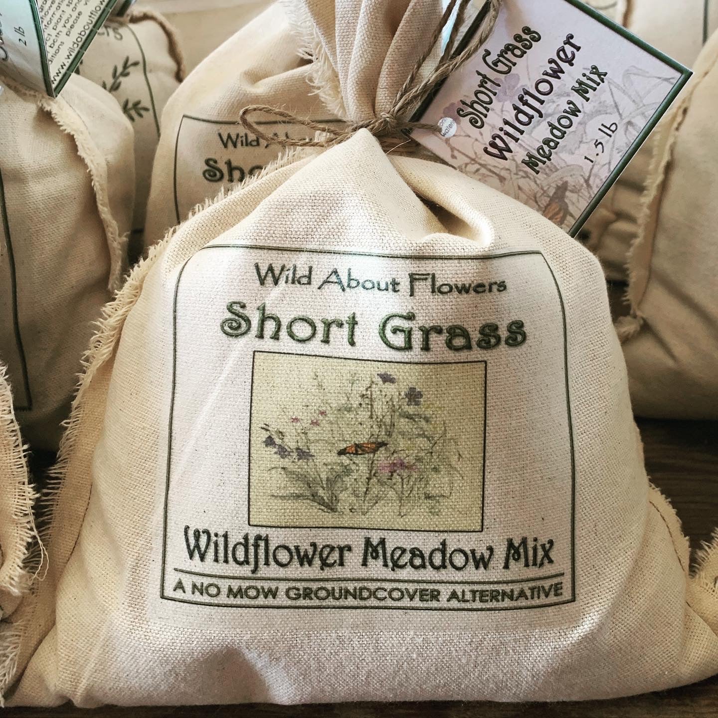 A cloth bag labeled "Wild About Flowers - Short Grass Wildflower Meadow Mix" sits in the foreground, tied at the top with twine and a product tag. The design features an illustration of a butterfly among wildflowers and grasses. Below the image, the bag reads "A No Mow Groundcover Alternative." Additional seed bags are visible in the background, contributing to a rustic and earthy display.